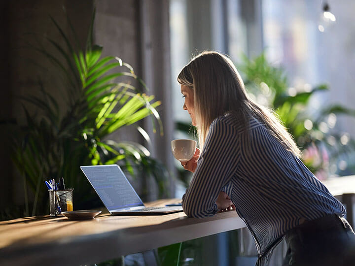 A woman sits looking a laptop screen while drinking coffee